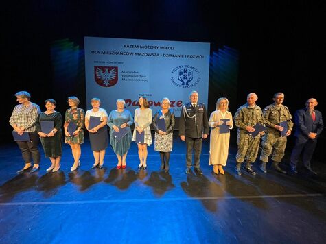 Dziesięcioro osób stoi na scenie w rzędzie i pozuje do zdjęcia. Wśród nich są dwaj żołnierze w strojach moro