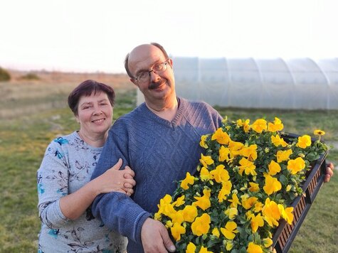 Uśmiechnięty mężczyzna w okularach trzyma skrzynkę pełną bratków. Obok niego stoi uśmiechnięta kobieta i trzyma mężczyznę pod ramię.