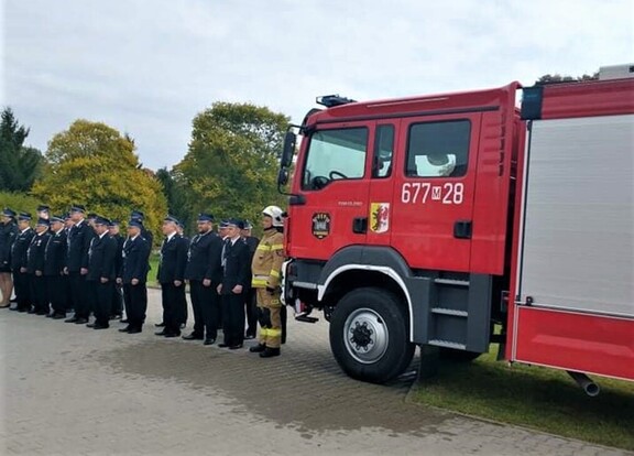 Przed wozem strażackim stoją druhowie ustawieni w jednym rzędzie. Pierwszy z nich jest w stroju ratowniczym