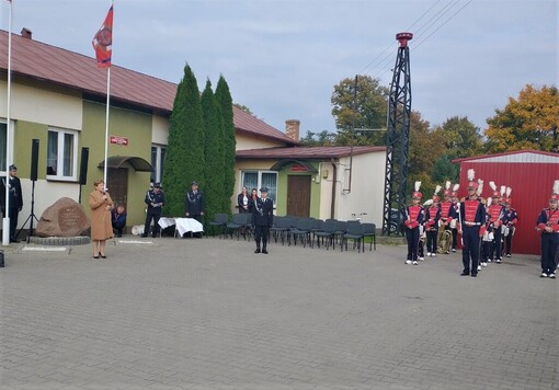 Na dziedzińcu przed remizą stoi kobieta z mikrofonem w prawej dłoni. Przed nią w znacznej odległości widać członków orkiestry strażackiej