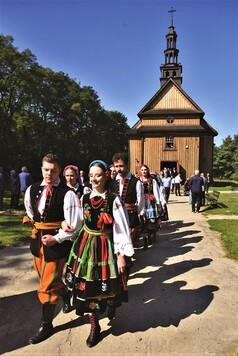 Korowód ludzi w strojach ludowych na tle drewnianego kościoła