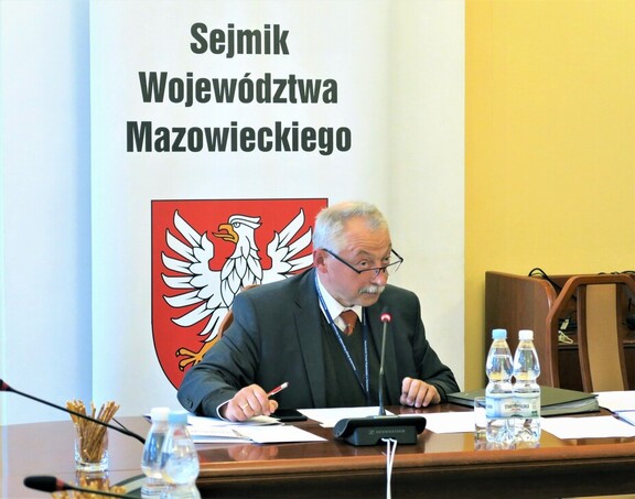 Starszy mężczyzna w garniturze i okularach siedzi przy stole konferencyjnym i mówi do mikrofonu. Brwi ma uniesione. Za nim jest baner Sejmiku i godło Mazowsza