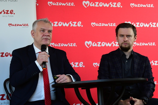 Dwóch mężczyzn siedzi przy stoliku na tle ścianki promocyjnej. Mężczyzna z lewej trzyma mikrofon.