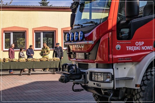 Czoło wozu strażackiego. W dali widać remizę i stół z rozłożonymi kompletami ubrań. Obok nich są strażacy