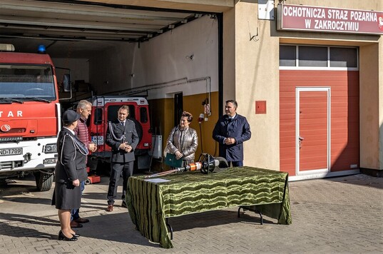 Kobieta stoi przed stołem z nożycami hydrauliczymi. Obok niej stoją trzej mężczyźni i kobieta w umundurowaniu strażackim