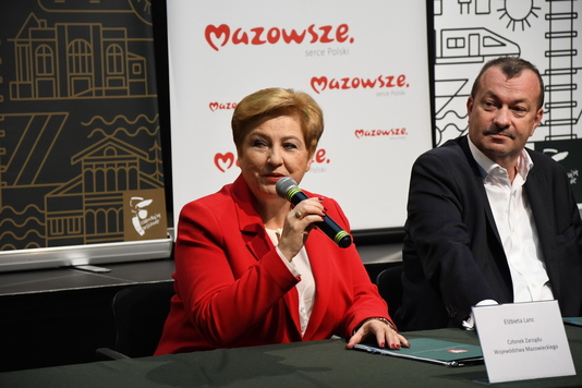 Elżbieta Lanc siedzi za stołem i przemawia do mikrofonu, który trzyma w prawej dłoni