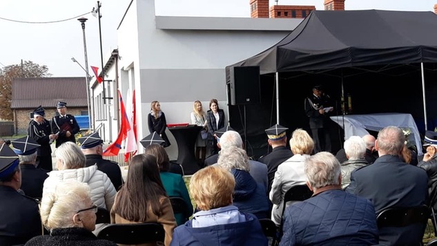 Podczas uroczystości. Na pierwszym planie na krzesłach siedzą zaproszeni goście. W tle widać oddany do użytku wyremontowany budynek świetlicy wiejskiej 