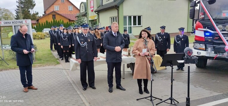 Janina Ewa Orzełowska stoi na placu przed remizą i przemawia do mikrofonu. Za nią stoi strażak i mężczyzna w garniturze