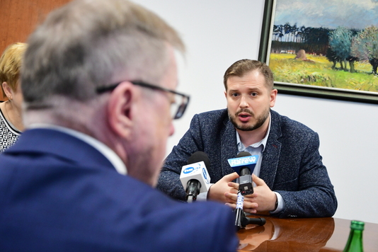 Elegancko ubrany mężczyzna siedzi przy stole i mówi do stojących przed nim mikrofonów.