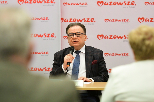 Marszałek siedzi za stołem i mówi do mikrofonu, który trzyma w prawej ręce