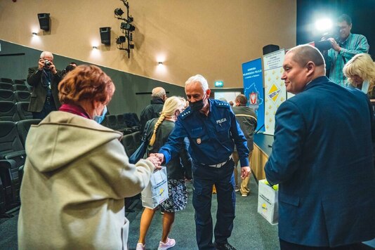 Policjant w mundurze wręcza upominek seniorce. W tle wnętrze sali widowiskowej.