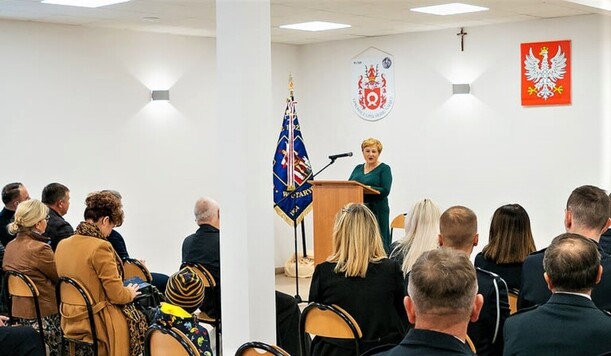 Kobieta w zielonej sukni na mównicy przed siedzącymi na krzesłach strazakami