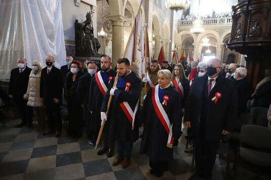 Samorządowcy przepasani biało-czerwonymi szarfami stoją wewnątrz kościoła. Wśród nich jest dyrektor Tomasz Kominek