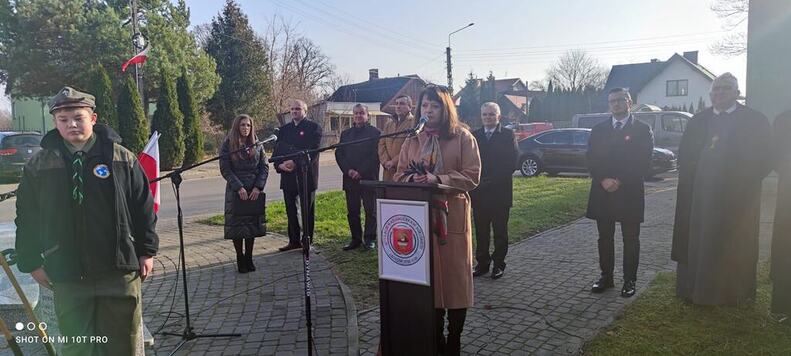 Janina Ewa Orzełowska stoi przy ustawionej na placu ambonie z herbem Cegłowa. Przemawia do mikrofonu. 