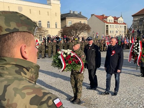 Wicemarszałek i radny Gołąbek idą obok siebie przez plac. Przed nimi kroczy żołnierz, który niesie wieniec