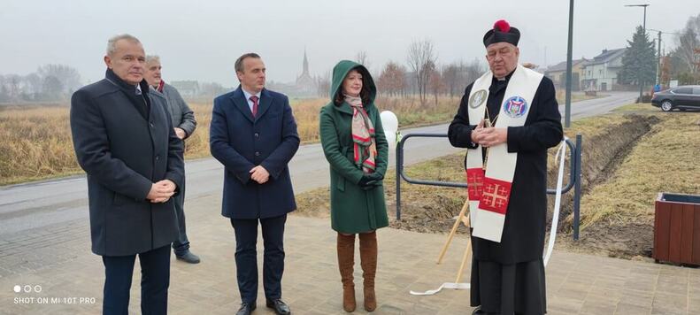 Janina Ewa Orzełowska, dwóch mężczyzn oraz ksiądz stoją na placu z siłownią