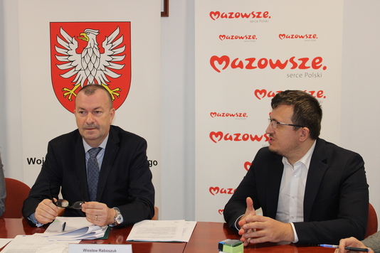 Mężczyźni siedzą przy stole, w tle logotypy Mazowsza. Przygotowuja sie do podpisania umów.