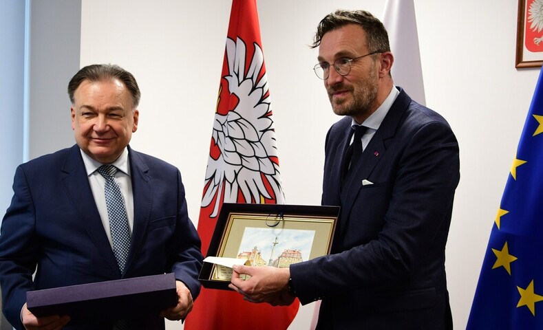 Marszałek i Sekretarz stanu regionu Bruksela-stolica stoją obok siebie na tle flag. Sekretarz stanu pokazuje podarowany mu obraz Starego Miasta w Warszawie. Marszałek tez trzyma w ręku pudełko