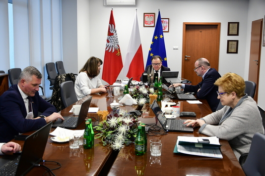 Grupa elegancko ubranych mężczyzn i kobiet siedzi przy stole, patrząc w ekrany komputerów. W tle flagi Mazowsza, Polski i Unii