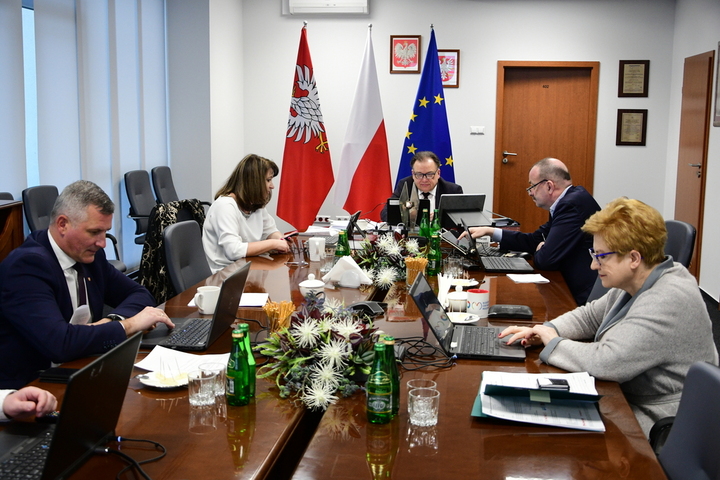 Grupa elegancko ubranych mężczyzn, kobiety i mężczyźni, siedzą przy stole i pracują przy komputerach. W tle flagi Mazowsza, Polski i Unii Europejskiej.