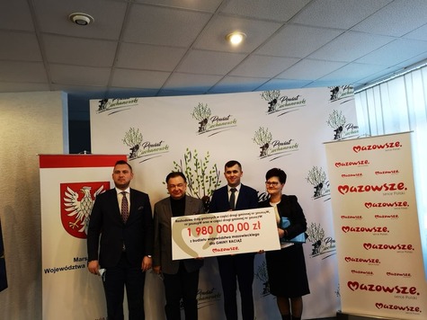 Grupa elegancko ubranych osób pozuje na tle emblematów związanych z Mazowszem, prezentując podpisany czek.