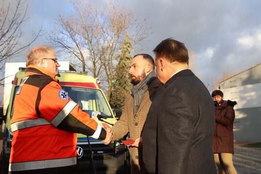 Elegancko ubrani mężczyźni ściskają dłoń ratownikowi medycznemu w pomarańczowym kombinezonie. W tle żółty ambulans.