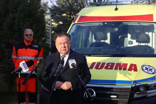 Elegancko ubrany mężczyzna mówi do mikrofonu. W tle żółty ambulans.