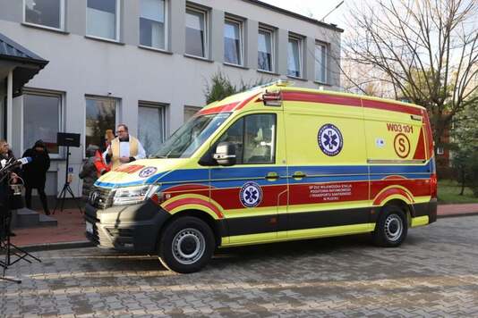 Ksiądz święci żółty ambulans, w tle ośrodek zdrowia.