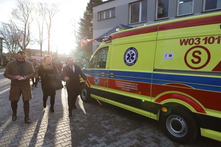 Dwóch mężczyzn i kobieta idą brukowanym parkingiem, mijają żółty ambulans.