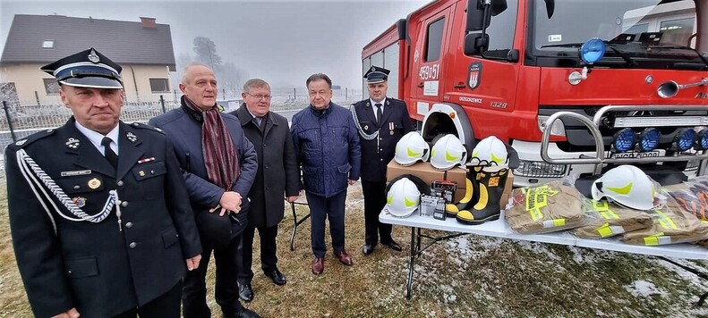 Marszałek i przewodniczący Przybytniak stoją wraz z dwoma strażakami i wójtem przy stole, na którym lezy zakupiony sprzęt. Obok widać wóz strażacki