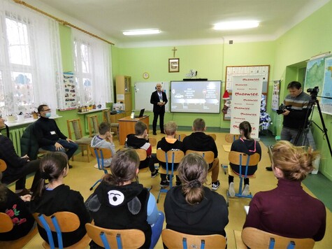 Przy tablicy z wyświetlaną prezentacją stoi prowadzący zajęcia. Przed nim na krzesłach siedzą uczniowie