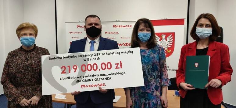 Grupa elegancko ubranych osób w maseczkach prezentuje czek z wypisaną kwotą dofinansowania. W tle emblematy Mazowsza.