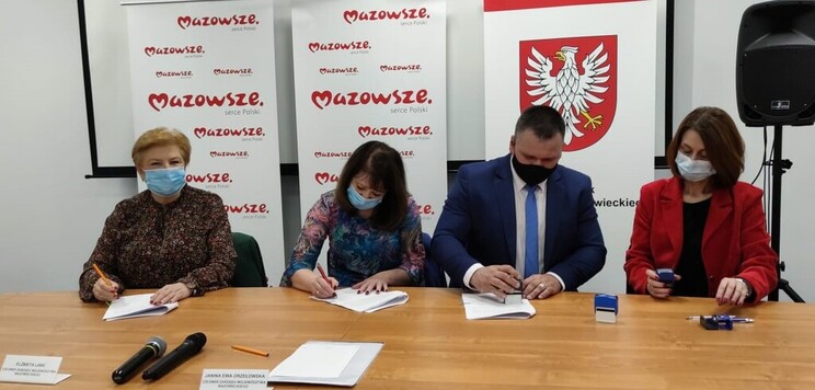 Grupa osób w maseczkach podpisuje dokumenty siedząc przy stole. W tle emblematy Mazowsza.