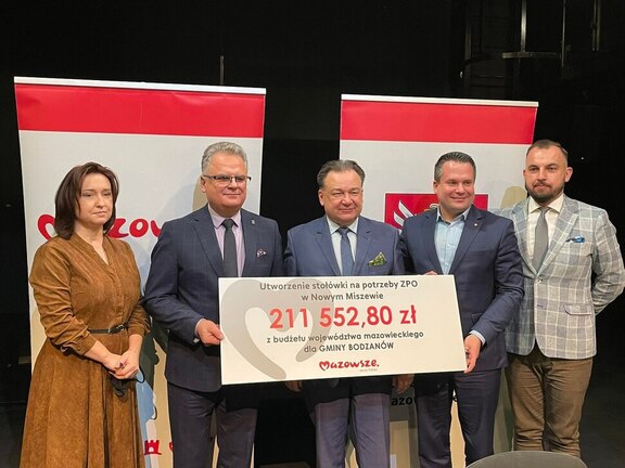 Pięć osób stoi na tle promocyjnych ścinkach. Trzymają biało czerwony duży czek