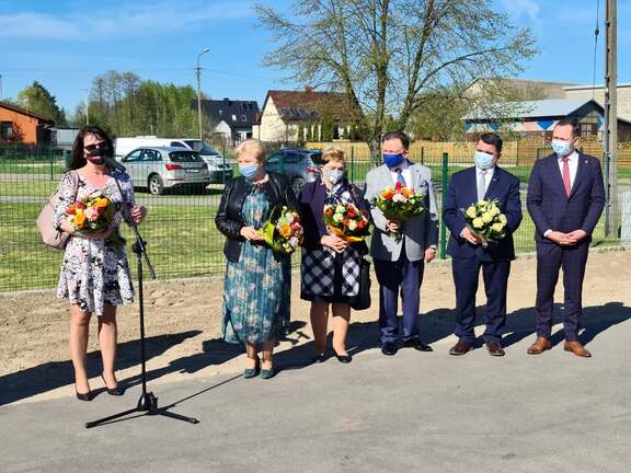 Marszałek Adam Struzik, Janina Ewa Orzełowska i Elżbieta Lanc stoją na zewnątrz wraz z trzema innymi osobami. Wrękach trzymają bukiety kwiatów. Janina Ewa Orzełowska stoi przed mikrofonem i przemawia.