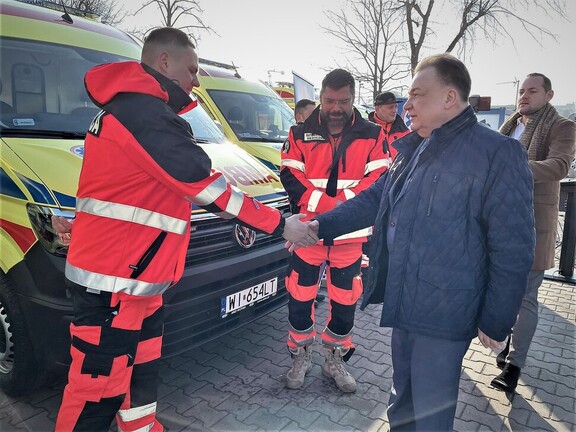 Marszałek i stojący naprzeciwko niego ratownik medyczny podają sobie ręce. W tle widać przekazane karetki