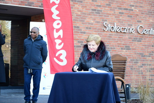 Członek zarządu województwa mazowieckiego siedzi przy stole i podpisuje umowę. W tle flaga Mazowsze