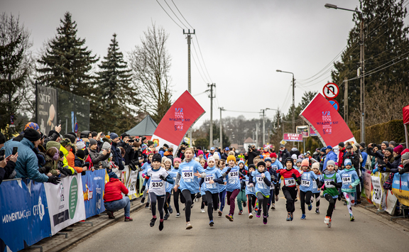 Dzieci w takich samych koszulkach biegną w maratonie. Zdjęcie z przodu grupy biegnących dzieci. Na poboczach ulicy stoją kibice