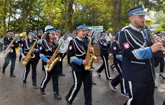 Muzycy z orkiestry dętej idą ulicą w pochodzie. każdy trzyma przed sobą instrument. Na pierwszym planie muzycy trzymają puzony