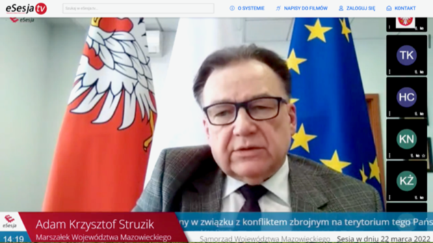 Stopklatka z transmisji sesji sejmiku online. Twarz i ramiona marszałka w pełnym planie, za nim flagi Mazowsza, Polski i Unii Europejskiej