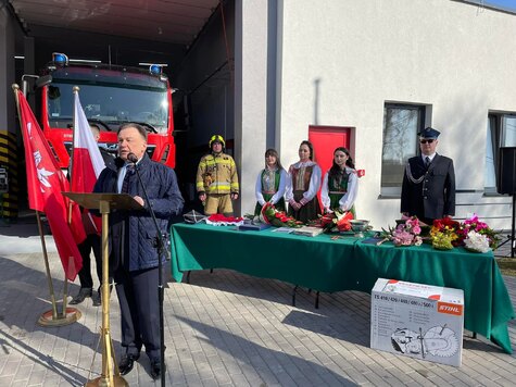 Marszałek stoi przy mównicy przed remizą z otwartym garażem. Mówi do mikrofonu