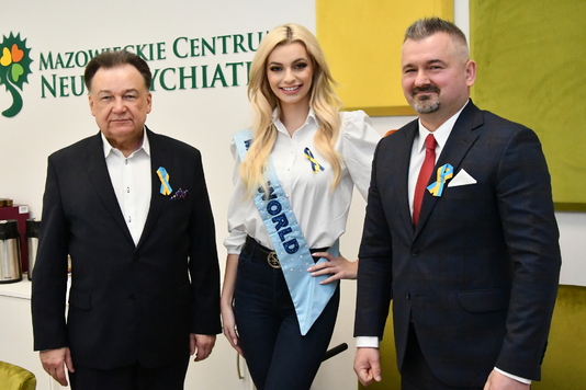 Kobieta w szarfie z napisem miss world i dwóch mężczyzn w garniturach stoją na tle ściany z napisem 