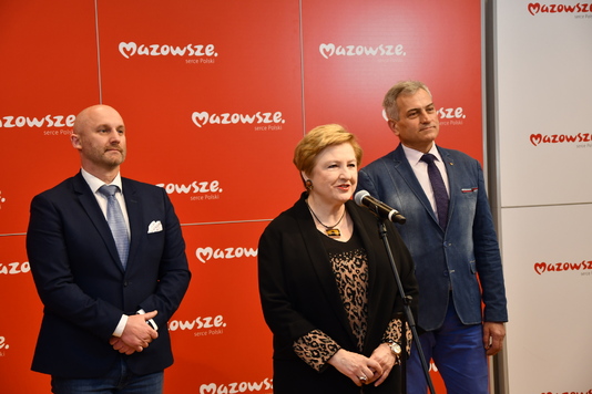 Kobieta z krótkimi włosami przed mikrofonem, obok dwóch mężczyzn elegancko ubranych, w tle ścianka promocyjna.