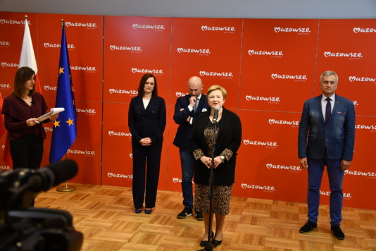 Konferencja prasowa, elegancko ubrani ludzie stoją na tle ścianki promocyjnej, jedna kobieta przemawia przez mikrofon.