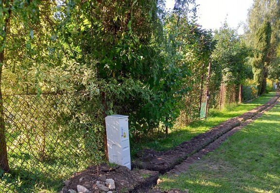 Rozkopana po jednej stronie aleja na terenie ogrodu działkowego. Obok rozkopanego wąskiego rowu stoi nowa skrzynka z rozdzielnią prądu