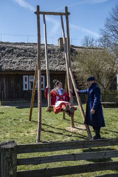 Przed wiejską chatą stoi wysoka drewniana huśtawka. Siedzi na niej dziewczyna w ludowym stroju, która patrzy na stojącego obok młodzieńca