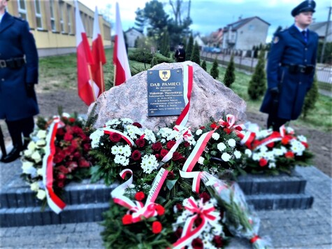 Wielki kamień, na którym jest tablica pamiątkowa. Koło pomnika stoją trzy flagi Polski. Przed pomnikiem leżą trzy wielkie wieńce