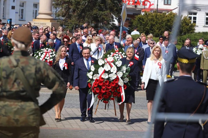 Marszałek oraz trzy kobiety idą obok siebie przez plac. Marszałek niesie, wraz z jedną z kobiet, wielki wieniec ułożony z białych i czerwonych kwiatów. Za nimi w tle widać innych uczestników uroczystości