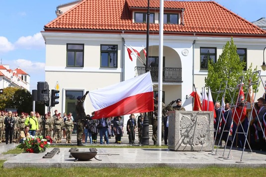 Na maszt wciągana jest wielka biało-czerwona flaga. Za masztem stoją w rzędzie harcerze