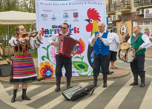 Trzech mężczyzn z harmonią, fletem i bębnem stoją obok siebie. Od lewa stoi kobieta w ludowym stroju. Gra na skrzypcach